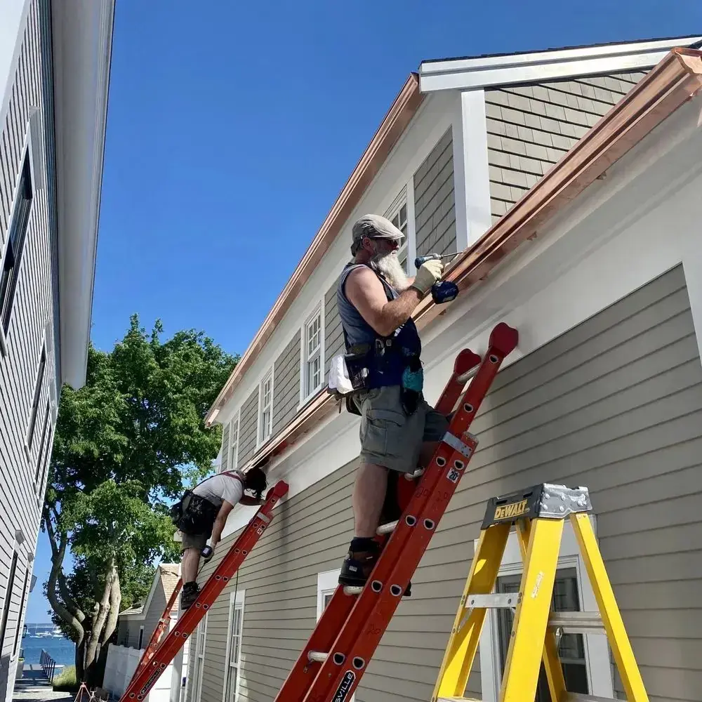 Installer on a ladder fastening a new copper gutter along the fascia of a gray shingled house